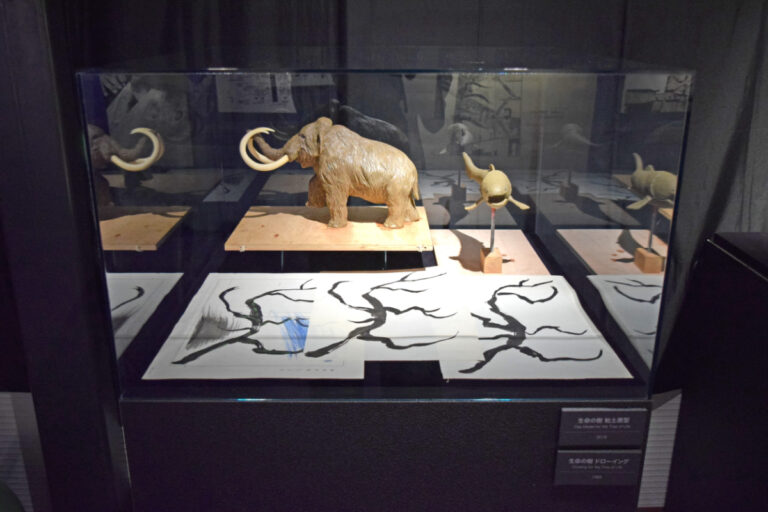 奥の生物は、1970年当時の写真をもとに作られた粘土原型（2018年）。手前の作品は、岡本による《生命の樹 ドローイング》（1968年）。